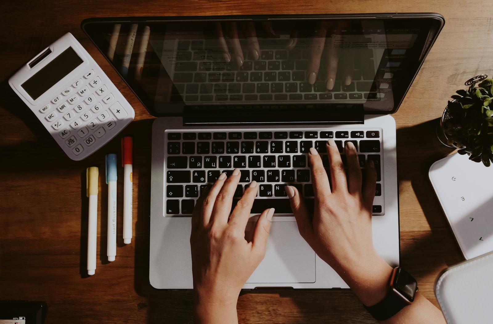 Hands typing on a laptop with a calculator and pens nearby on a wooden table.
